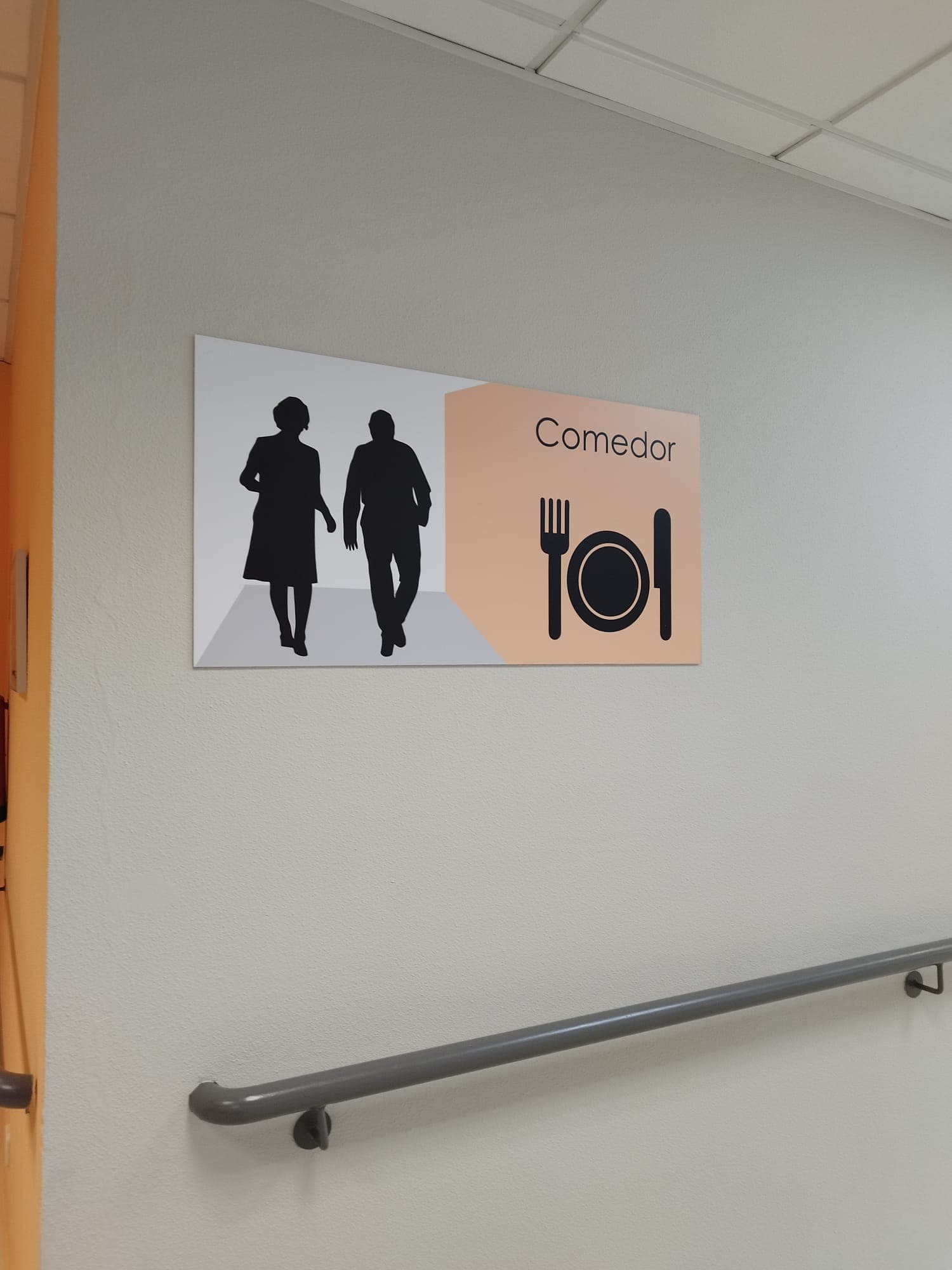 Señalización de comedor en centro para mayores. Cartel con siluetas caminando y símbolo de cubiertos. Barandilla en pared para apoyo. Diseño facilita orientación y movilidad segura.